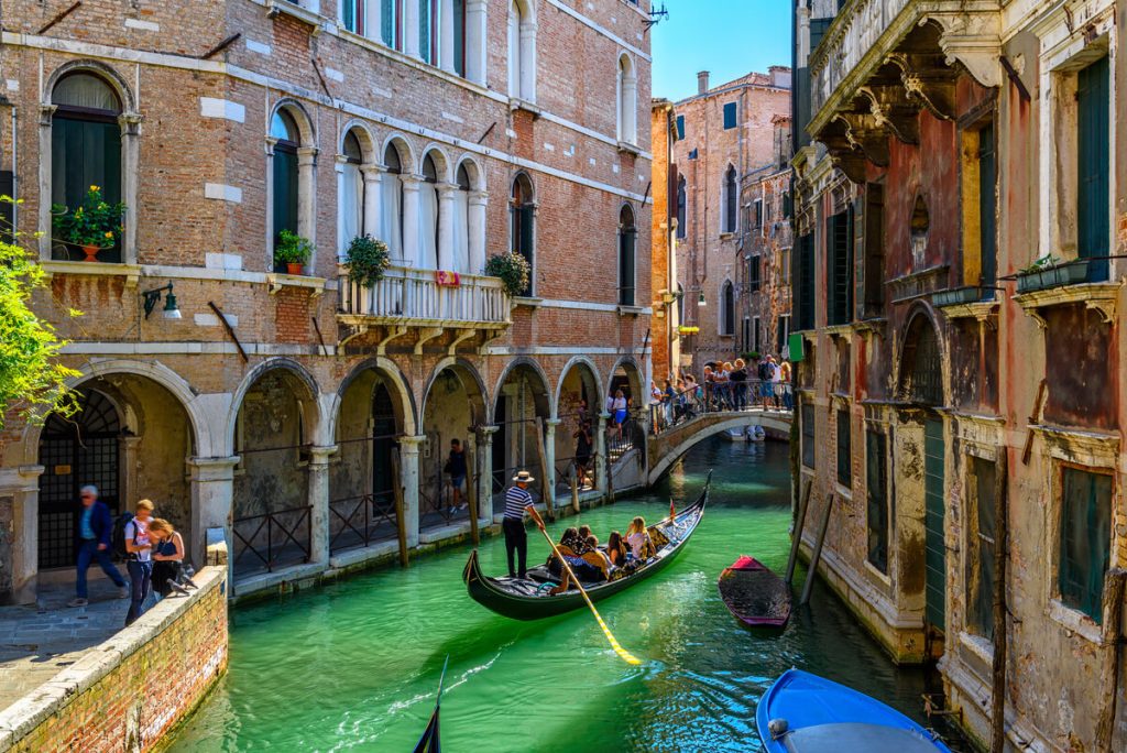 A gondolier rows passengers along a narrow Venice canal beneath a small pedestrian bridge