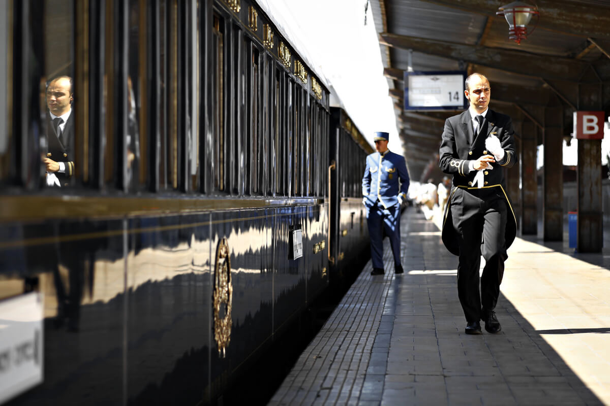 Uniformed attendants beside a blue Venice Simplon-Orient-Express carriage at a railway platform